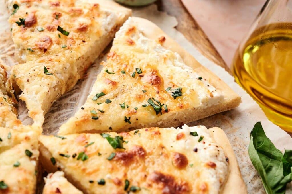 Sliced white pizza with melted cheese and herbs on a wooden board, next to a glass container of olive oil and fresh basil leaves, showcasing the classic flavors of white pizza.