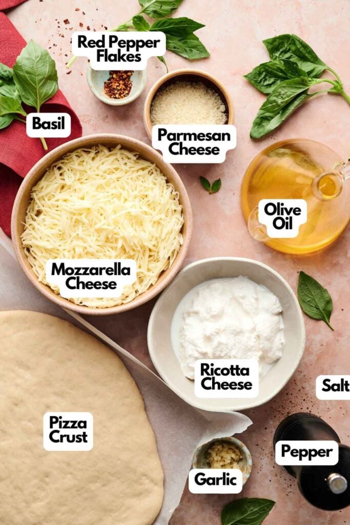 Ingredients for making White Pizza, including pizza crust, mozzarella cheese, ricotta cheese, parmesan cheese, basil, red pepper flakes, olive oil, salt, pepper, and garlic on a counter.