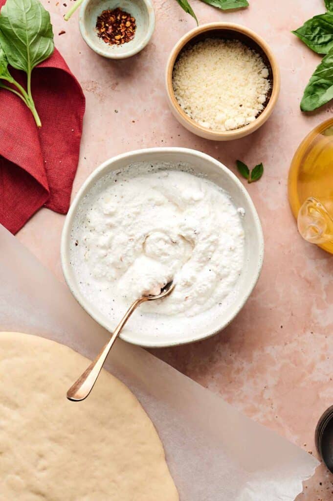 A bowl of white ricotta cheese spread with a spoon sits beside grated cheese, fresh spinach, red pepper flakes, olive oil, and pizza dough on parchment paper—perfect ingredients for making a delicious White Pizza.