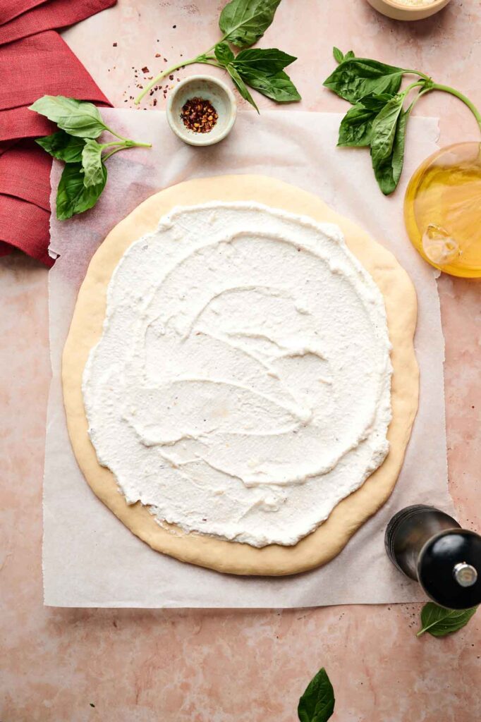 Unbaked White Pizza dough topped with creamy ricotta cheese sits on parchment paper, surrounded by basil leaves, olive oil, crushed red pepper, and a red napkin.