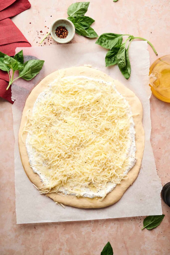 White Pizza topped with ricotta spread, shredded cheese, and black pepper on a round dough base, surrounded by fresh basil, olive oil, and a small bowl of red pepper flakes.