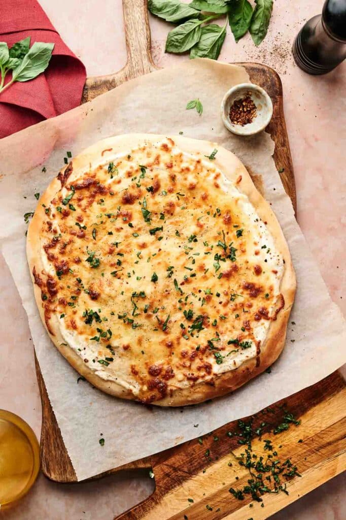 A baked white pizza topped with chopped herbs sits on parchment paper over a wooden board, with a small bowl of red pepper flakes and fresh basil nearby.