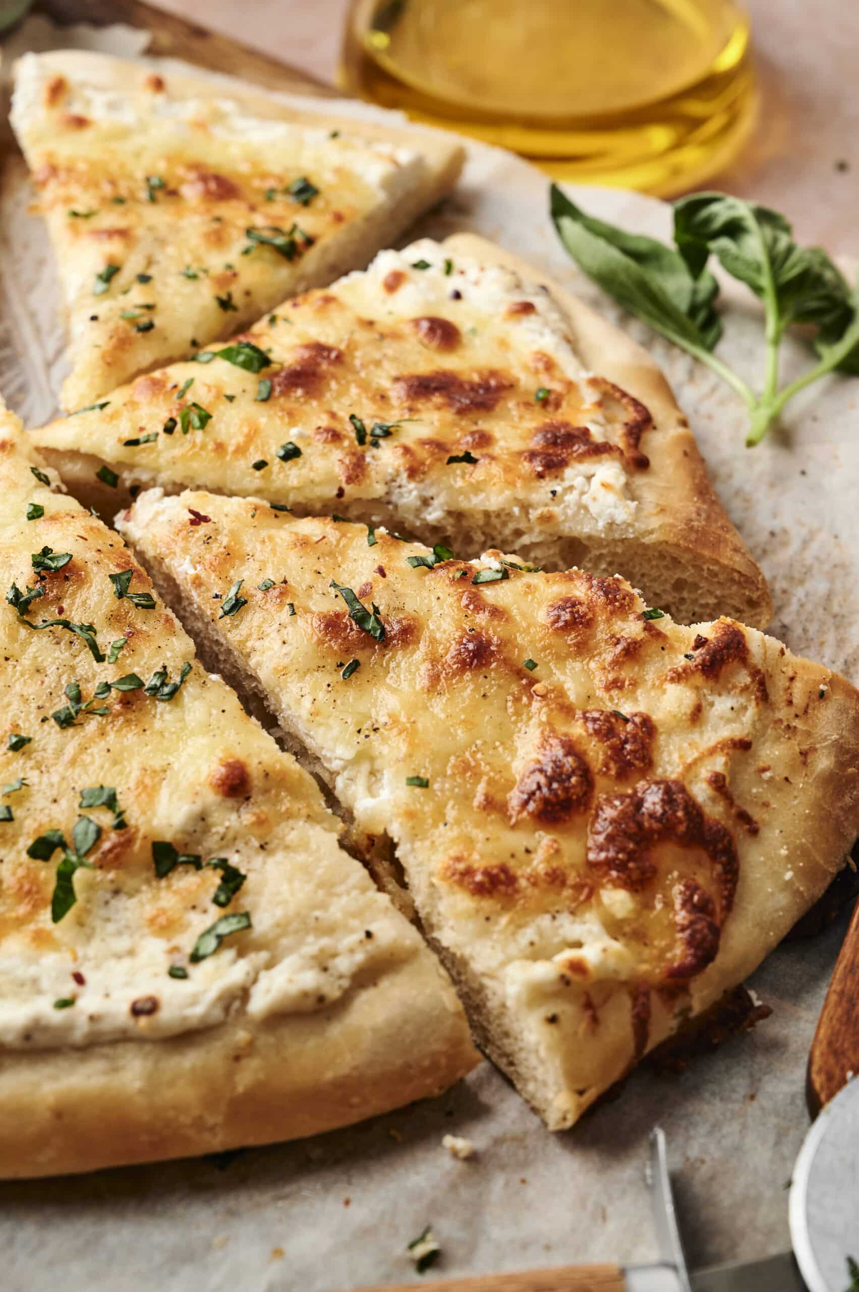 Slices of cheesy garlic bread with herbs, reminiscent of white pizza, rest on parchment paper, alongside a sprig of fresh basil and a glass container of olive oil.