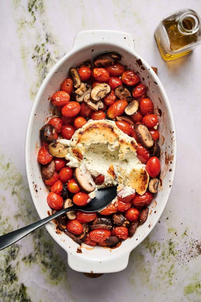 An oval baking dish with roasted cherry tomatoes, mushrooms, and a partially scooped baked cheese center, with a spoon resting inside and a small bottle of olive oil nearby.