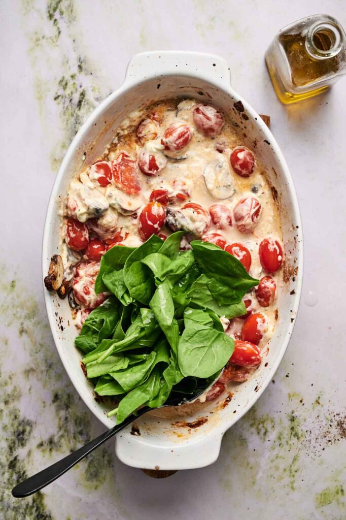 A white baking dish with roasted cherry tomatoes and creamy sauce, topped with fresh spinach leaves; a bottle of olive oil is nearby.
