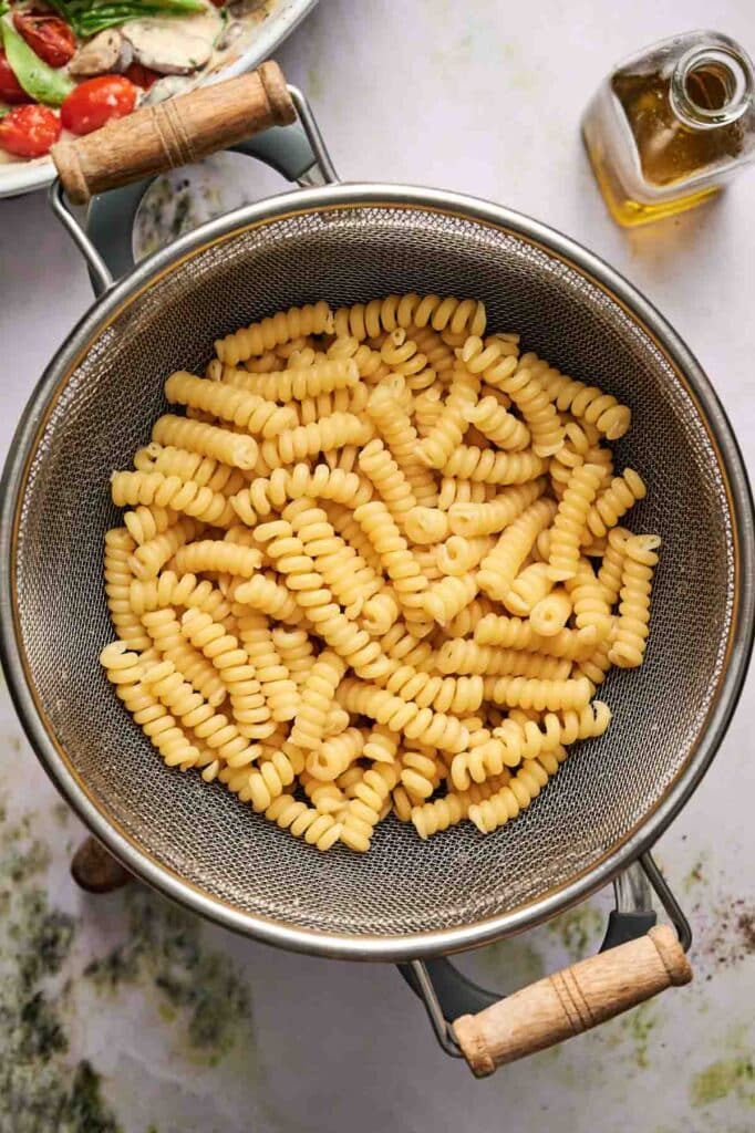 Cooked rotini pasta draining in a metal colander with wooden handles, with a small bottle of olive oil and a salad partially visible in the background.