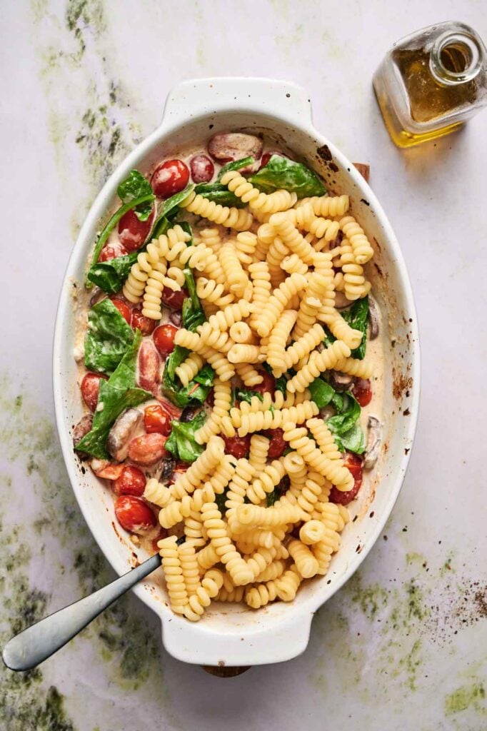 Oval baking dish with fusilli pasta, cherry tomatoes, spinach, and creamy sauce, with a metal spoon and a bottle of olive oil nearby.