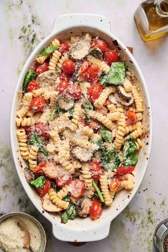 A white oval baking dish filled with fusilli pasta, cherry tomatoes, spinach, mushrooms, and topped with grated cheese. Olive oil and grated cheese jars are nearby.