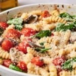 A white bowl filled with fusilli pasta, cherry tomatoes, mushrooms, spinach, and grated cheese. A bottle of olive oil is visible in the background.