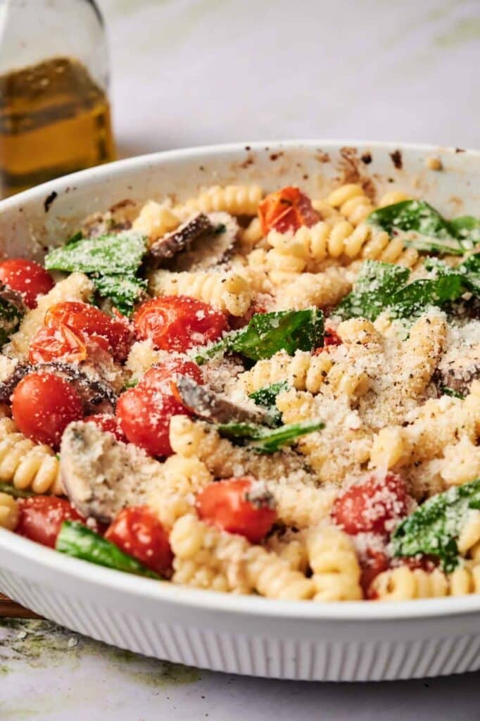 A white bowl of rotini pasta with cherry tomatoes, spinach, mushrooms, and grated parmesan cheese, with a bottle of olive oil in the background.