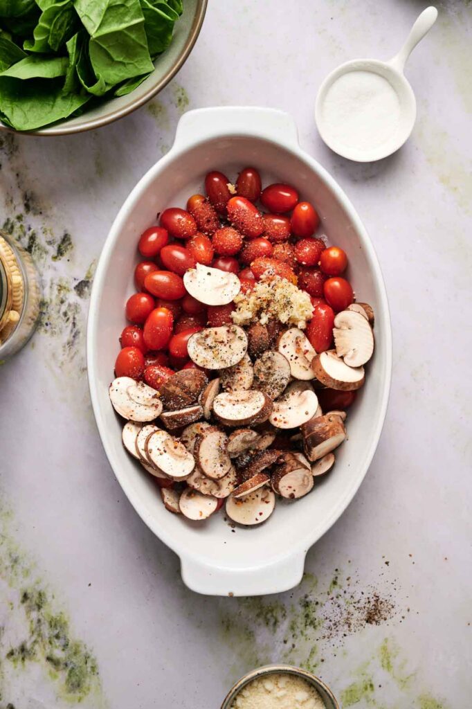 Oval baking dish filled with sliced mushrooms, cherry tomatoes, minced garlic, and cracked black pepper. Bowls of spinach and grated cheese are nearby on a marble surface.