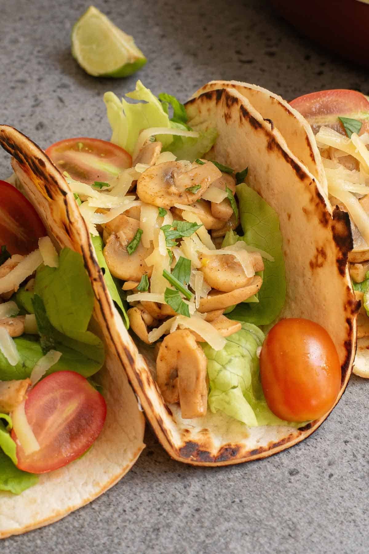 Two mushroom tacos filled with lettuce, sliced mushrooms, shredded cheese, tomato halves, and herbs rest on a gray surface, with a lime wedge in the background.