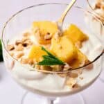 A glass dessert dish with fluffy yoghurt, pineapple chunks, chopped nuts, and a mint leaf garnish, with a spoon inserted into the creamy treat.