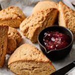 Several scones are arranged on parchment paper next to a small black bowl filled with red fruit jam.