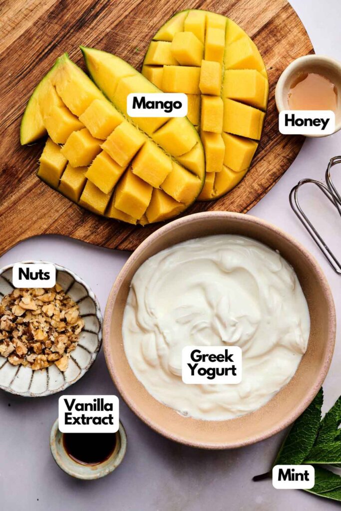 Sliced mango, fluffy yoghurt, chopped nuts, honey, vanilla extract, and mint are arranged on a table with labels identifying each ingredient.