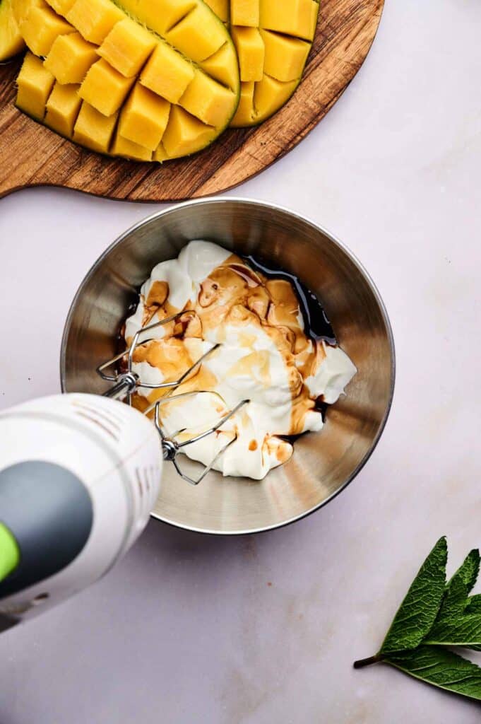 A hand mixer hovers above a metal bowl of fluffy yoghurt and syrup, with diced mango on a wooden board nearby and a green leaf resting on the surface.