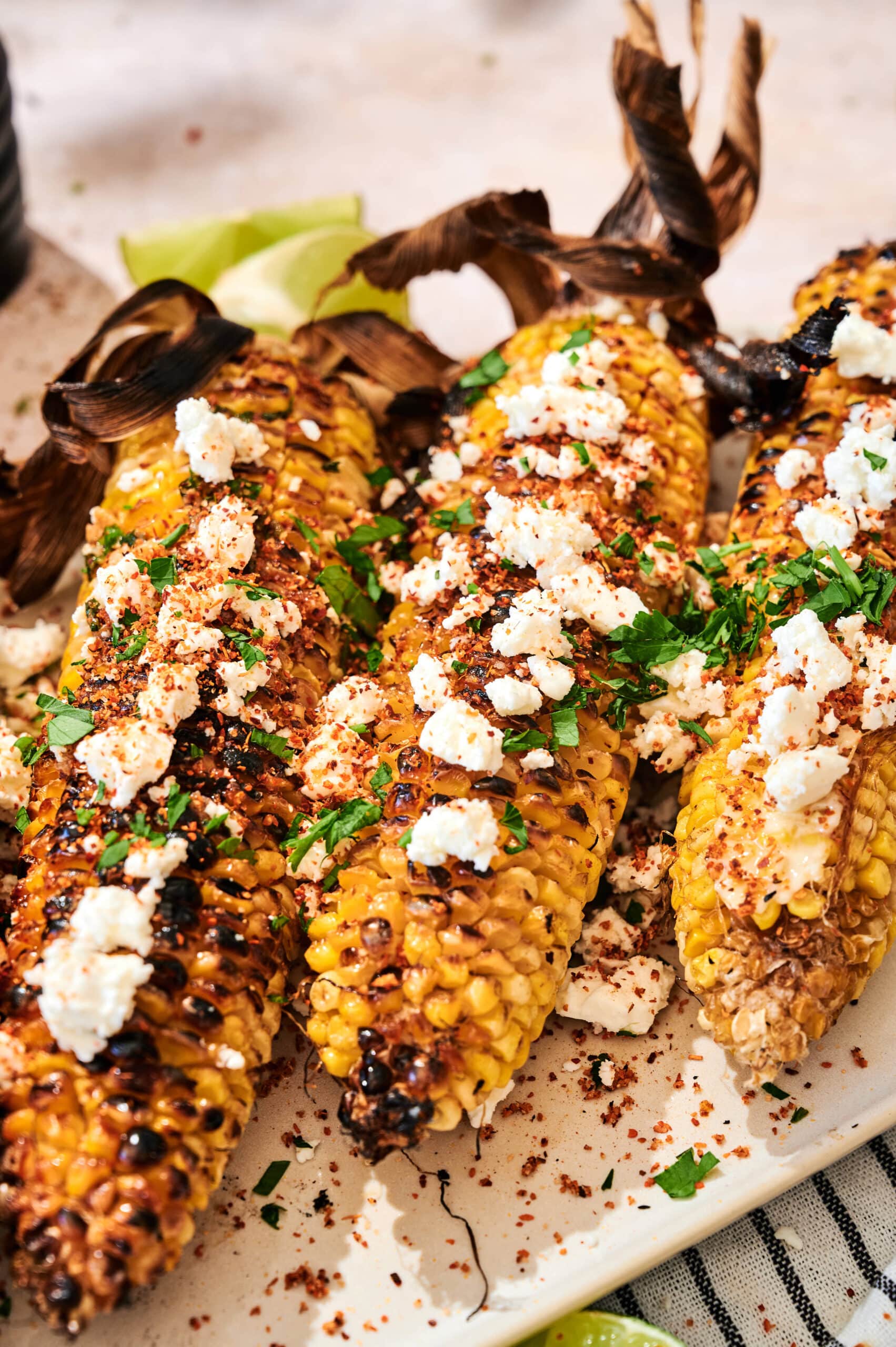 Four grilled corn cobs topped with crumbled cheese, chopped herbs, and chili powder are arranged on a plate.