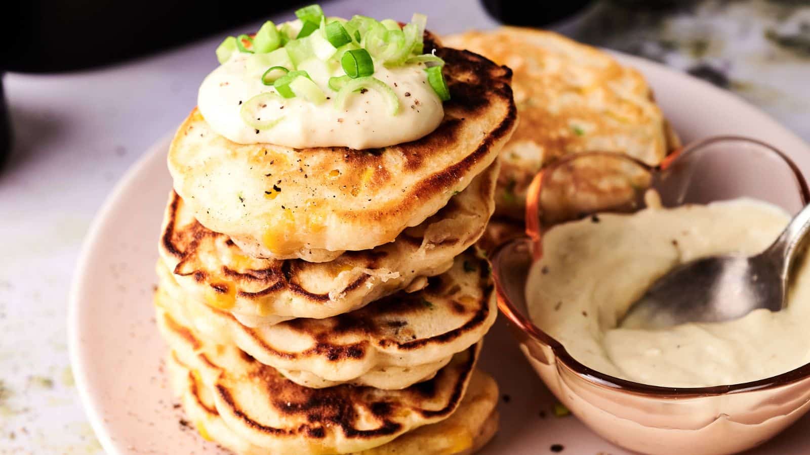 A stack of savory pancakes topped with sour cream and chopped green onions next to a bowl of creamy sauce with a spoon.