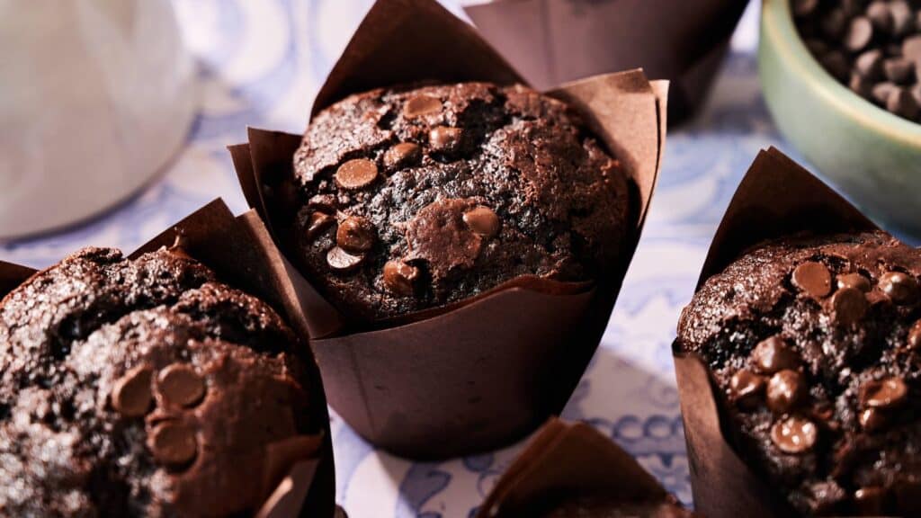 Close-up of chocolate muffins in brown paper wrappers, topped with chocolate chips, on a blue and white patterned surface.