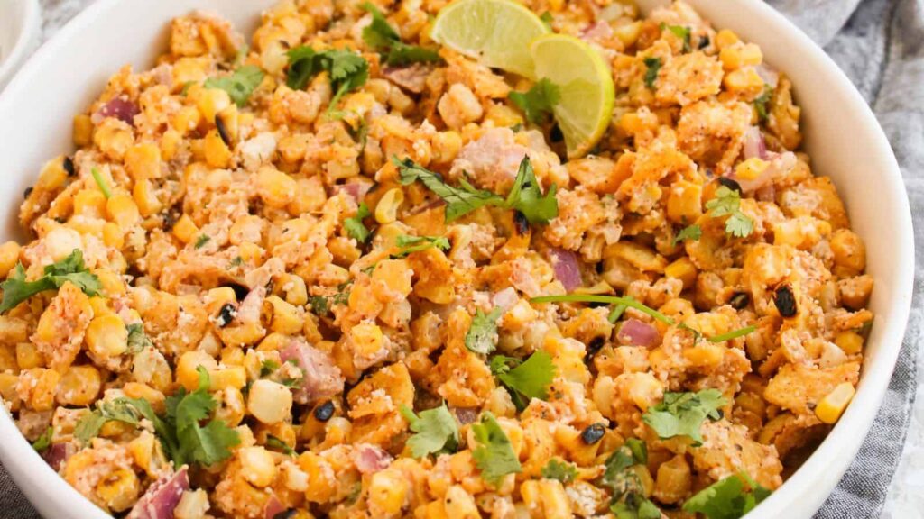 A bowl of Mexican street corn salad topped with fresh cilantro and lime wedges, featuring grilled corn, onions, cheese, and spices.