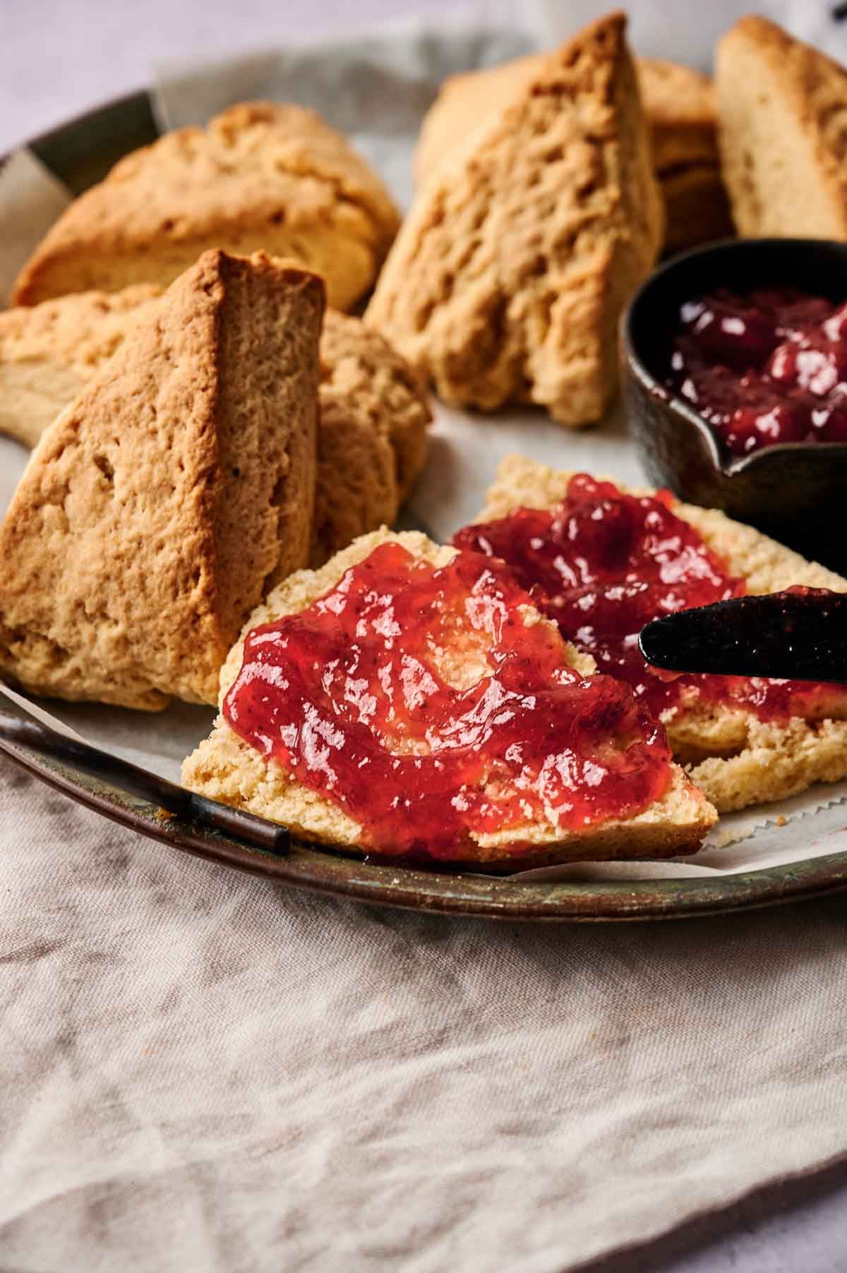A plate of scones, some whole and some split open, sits invitingly with one scone spread with red fruit jam and a butter knife resting on the plate.