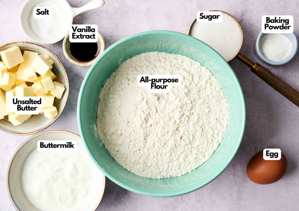 Various labeled baking ingredients for scones are arranged on a table, including all-purpose flour, sugar, baking powder, salt, unsalted butter, buttermilk, vanilla extract, and an egg.