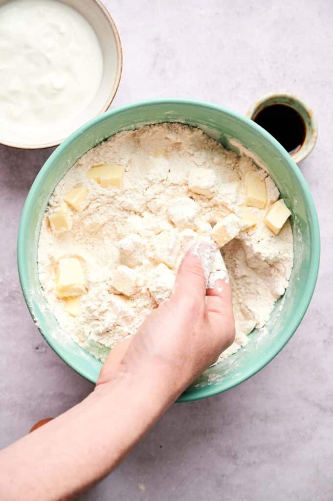 A hand mixes butter cubes into flour in a green bowl, preparing dough for scones, with a small bowl of yogurt and a cup of dark liquid nearby.