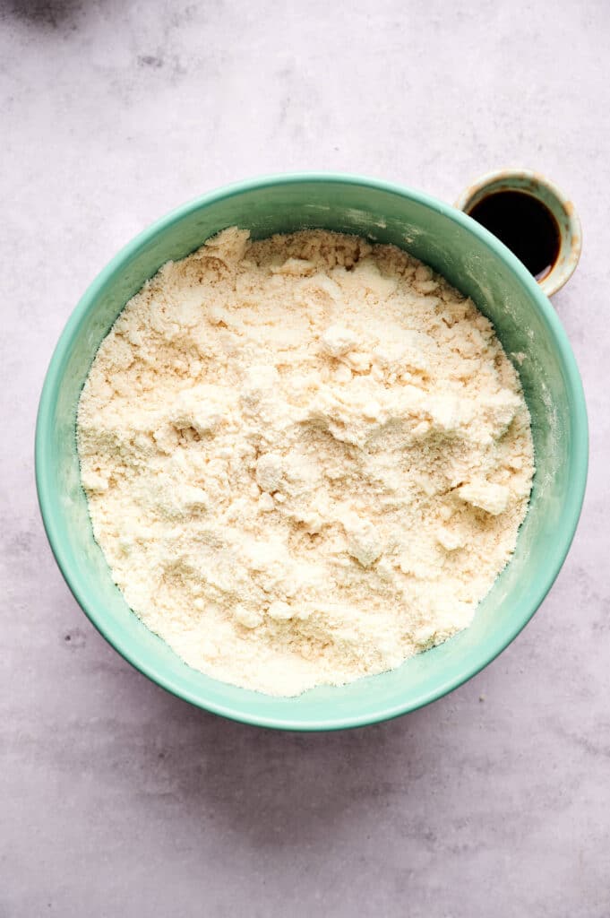 A green bowl filled with a light-colored, crumbly flour mixture for scones sits on a gray surface. A small cup with a dark liquid is partly visible beside the bowl.