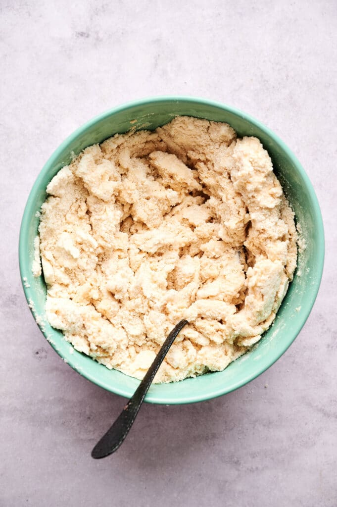 A light green bowl filled with a coarse, crumbly scone dough mixture and a metal spoon resting inside.