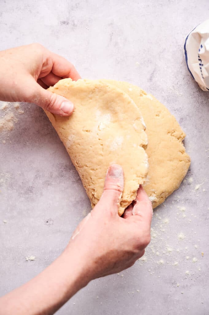 Two hands folding a piece of raw scone dough in half on a lightly floured surface.
