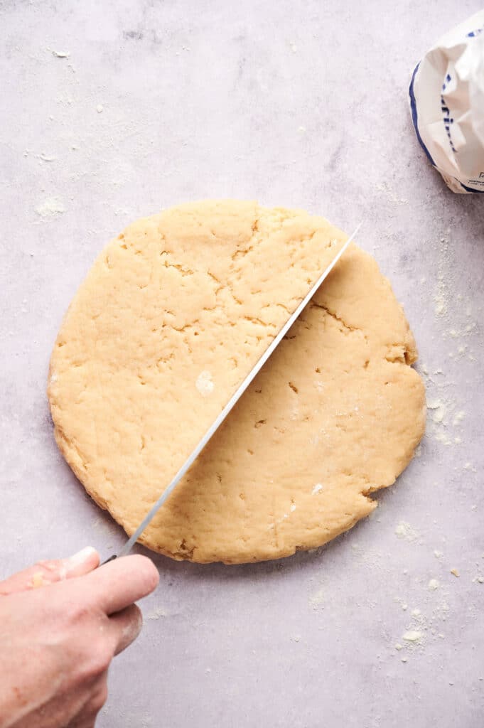A round disc of scone dough is being cut in half with a large knife on a floured surface.
