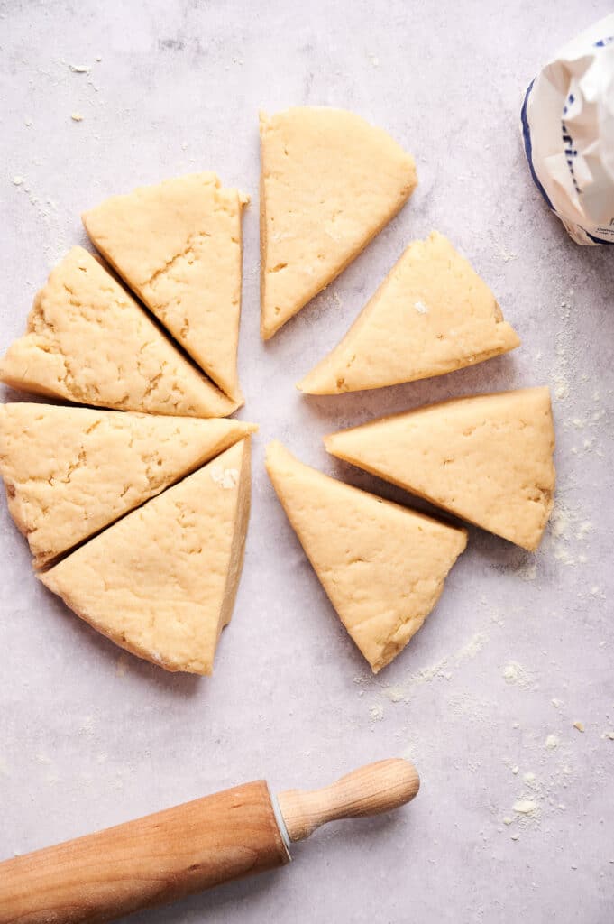 A round disc of scone dough, cut into eight triangular scones, sits on a floured surface with a rolling pin and a bag in view.