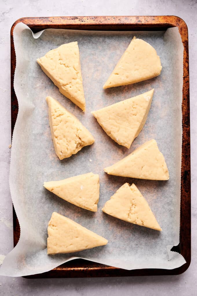 Eight unbaked, triangular scone dough pieces are arranged on a parchment-lined baking sheet, ready to be baked into delicious scones.
