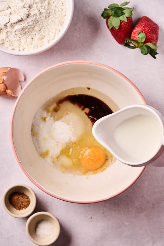 A mixing bowl with an egg, sugar, vanilla, and milk being poured in—surrounded by flour, strawberries, eggshells, and small bowls of spices and salt—sets the scene for making delicious homemade strawberry bread.