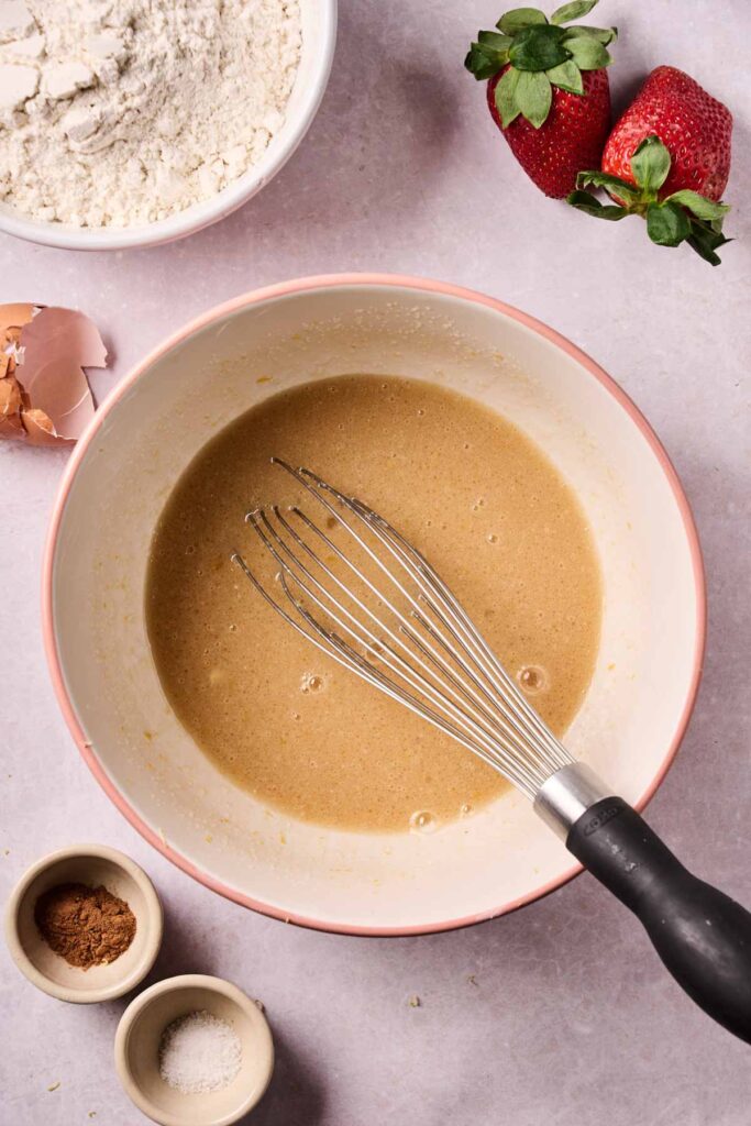 Bowl of mixed batter for strawberry bread with a whisk, surrounded by flour, strawberries, eggshell, and small bowls of spices and salt on a countertop.