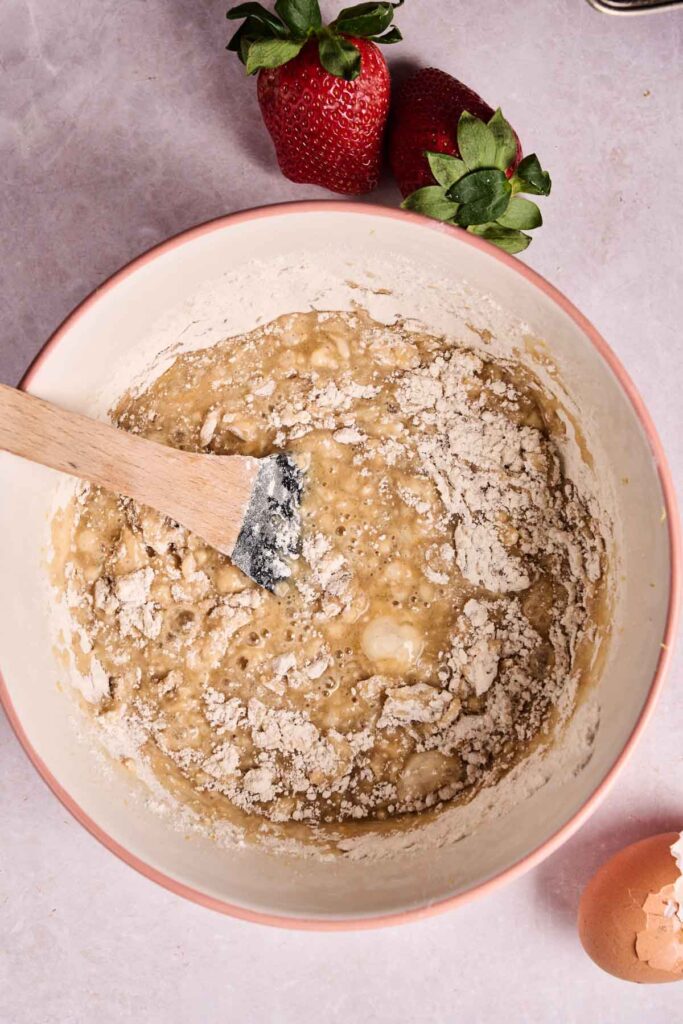 A mixing bowl with partially mixed strawberry bread batter, a wooden spatula, two strawberries, and a cracked eggshell rest on a light countertop.