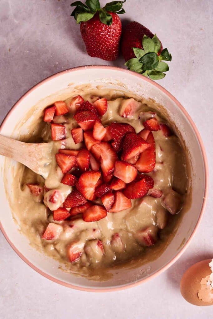 A mixing bowl with strawberry bread batter and chopped fresh strawberries, a wooden spoon, two whole strawberries, and a cracked eggshell nearby.