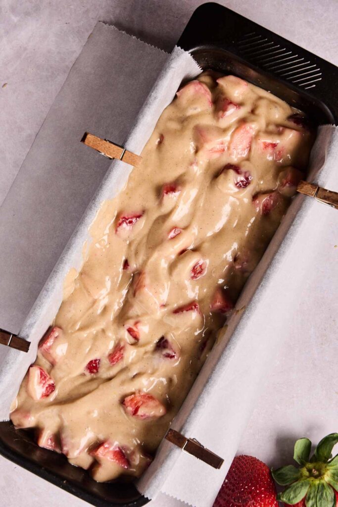 A loaf pan lined with parchment paper holds unbaked strawberry bread batter, with fresh strawberry pieces mixed throughout.