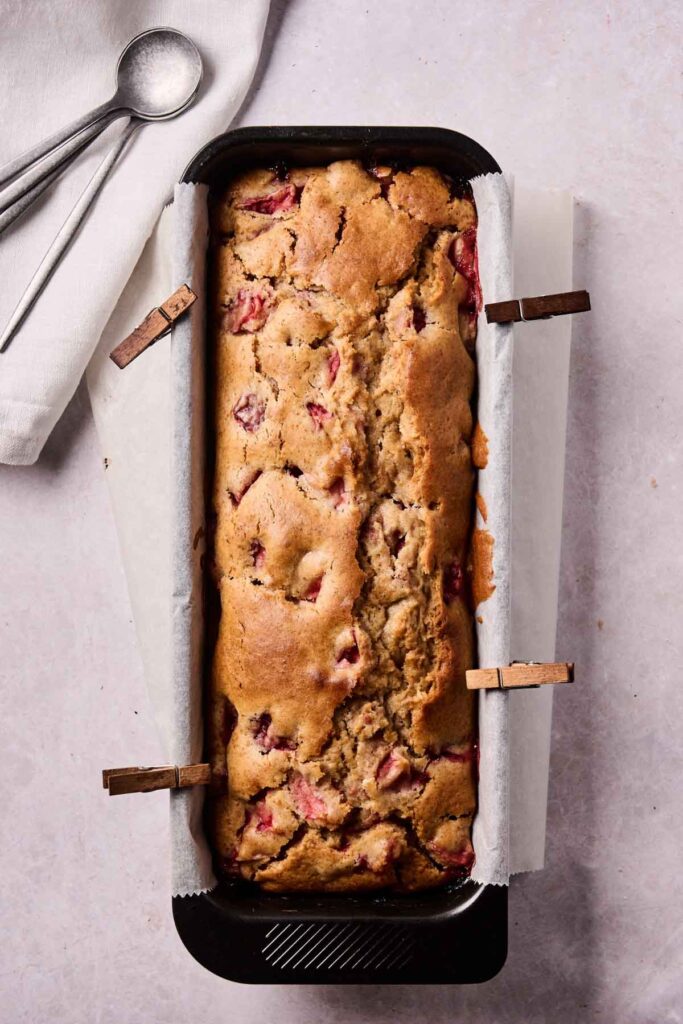 A baked loaf of strawberry bread in a parchment-lined pan, secured with clips, sits on a light surface beside a white napkin and two spoons&mdash;ready to enjoy this sweet strawberry bread treat.