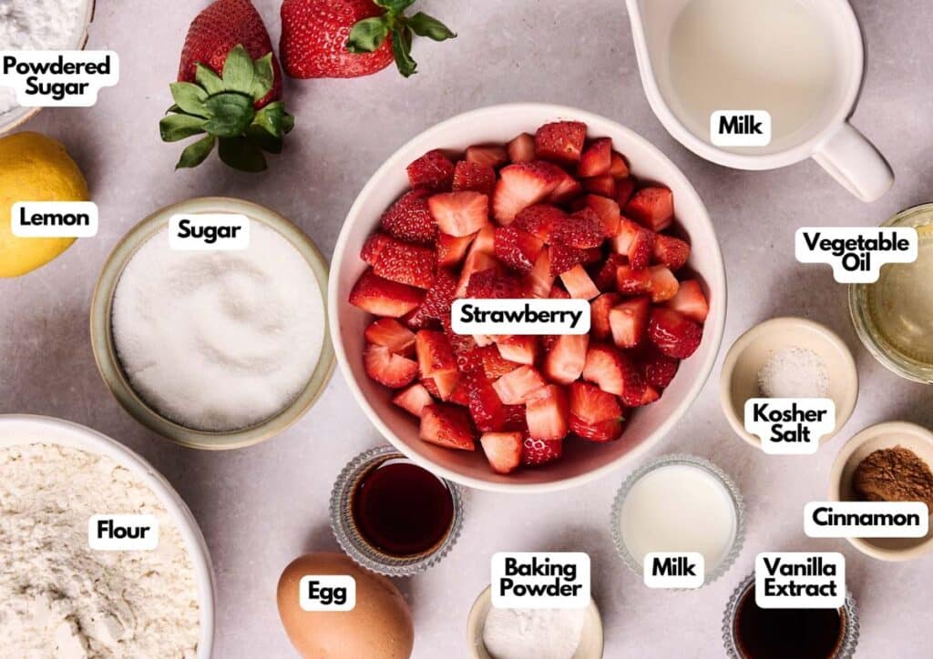 A top view of labeled baking ingredients for strawberry bread, including strawberries, sugar, lemon, flour, egg, milk, vanilla extract, cinnamon, vegetable oil, baking powder, kosher salt, and powdered sugar.