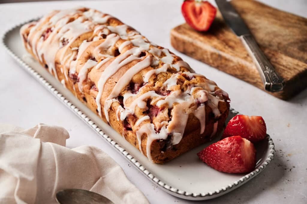 A rectangular loaf of strawberry bread with white icing drizzle sits on a platter, accompanied by three sliced strawberries. A knife rests on a wooden board in the background, completing the inviting strawberry bread display.