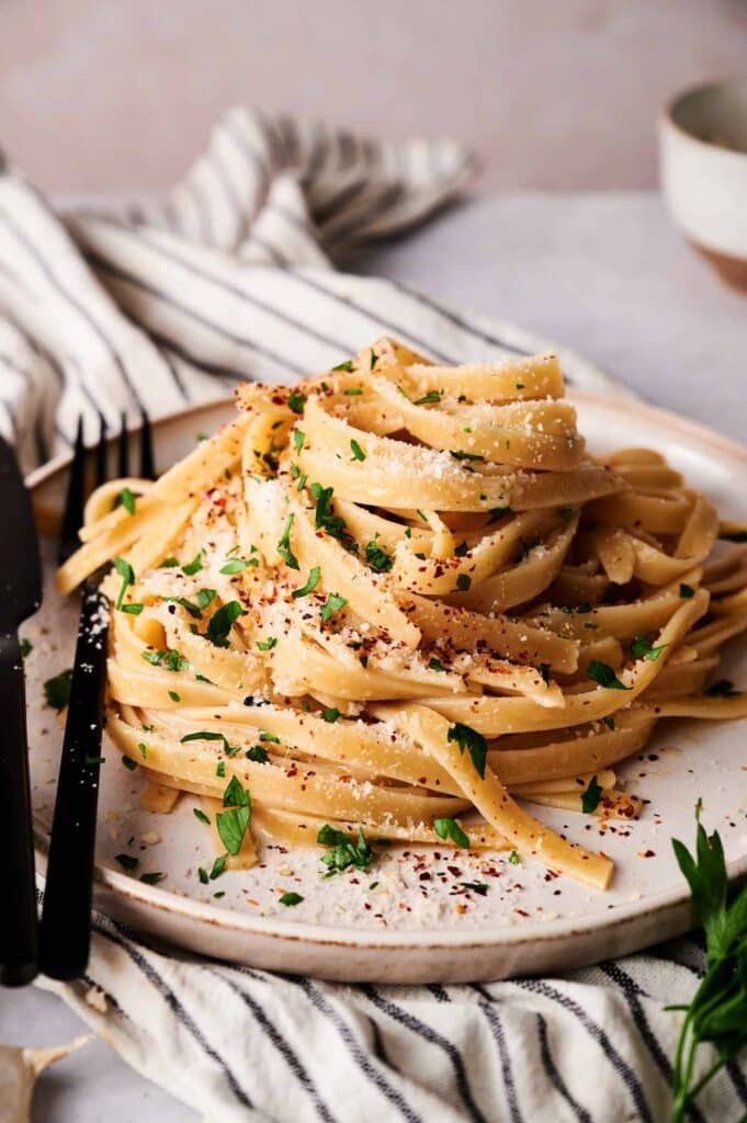 A plate of linguini aglio e olio topped with grated Parmesan, chopped parsley, and red pepper flakes, sits beside a fork and knife on a striped napkin.