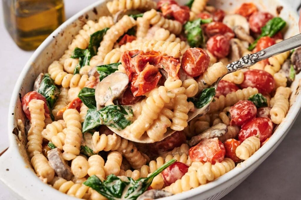 Boursin Cheese Pasta with a creamy sauce, cherry tomatoes, spinach, and mushrooms in a white baking dish, with a serving spoon lifting a portion.