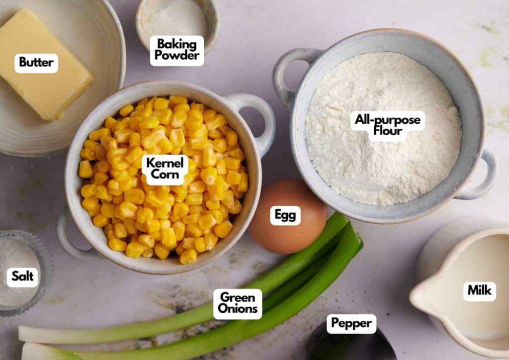 Top-down view of ingredients labeled kernel corn, all-purpose flour, egg, green onions, milk, pepper, salt, baking powder, and butter, arranged on a light surface.