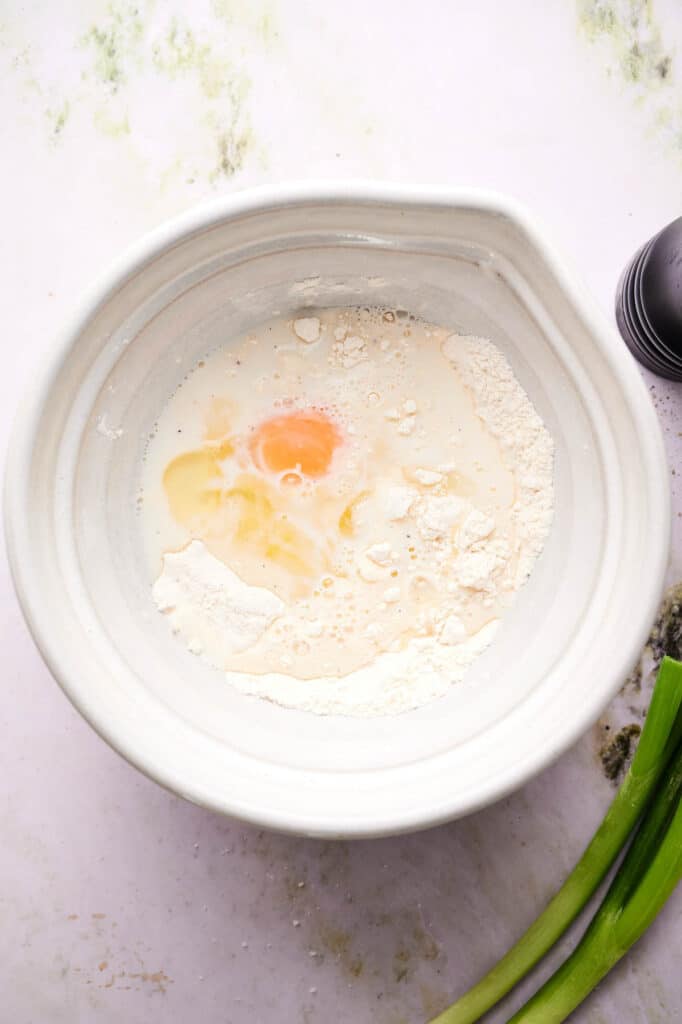 A white bowl containing flour, milk, and a cracked egg, with a black pepper grinder and green onions beside it on a light surface.