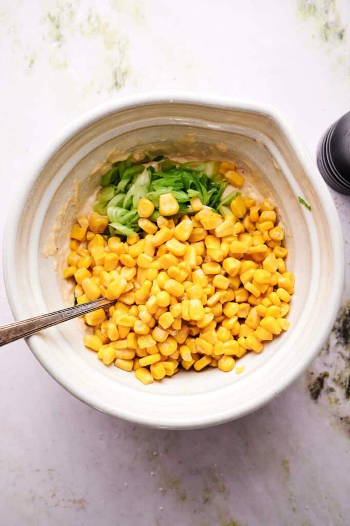 A white bowl contains corn kernels, chopped green onions, and a creamy mixture being stirred with a metal spoon.