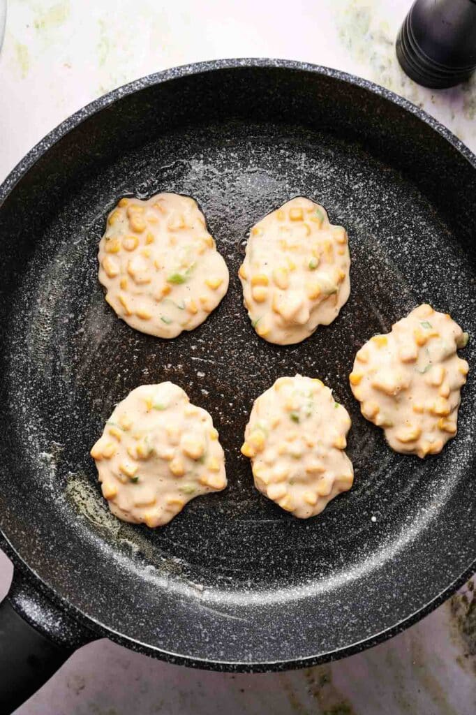 Five uncooked corn fritter patties with chopped green onions are spread out in a nonstick frying pan, ready to be cooked.