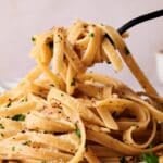 Close-up of a fork lifting linguini aglio e olio garnished with grated Parmesan, chopped parsley, and red pepper flakes.
