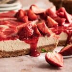 A sliced no bake strawberry cheesecake with a graham cracker crust, topped with strawberry sauce and fresh strawberries, sits on a light-colored surface.