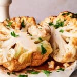 Two roasted whole cauliflower halves garnished with fresh chopped parsley are displayed on parchment paper.
